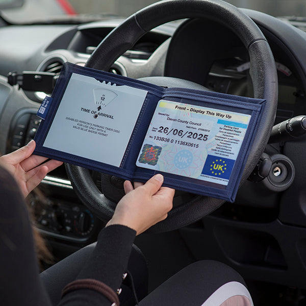 Open Disabled Blue Badge Wallet held in two hands against a steering wheel. The adjustable timer display clock is on the left. On the right is a Devon County Council Disabled Parking Permit with hologram. The acetate window doesn't cover the hologram.