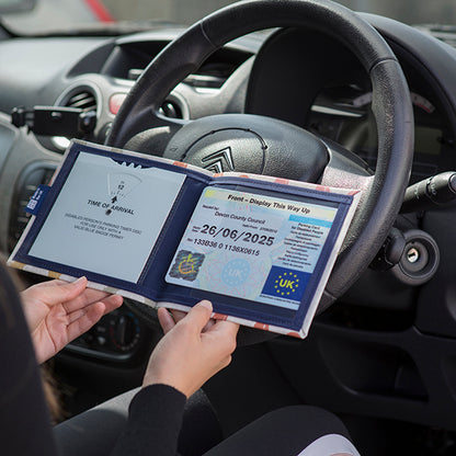 Open Disabled Blue Badge Wallet held in two hands against a steering wheel. The adjustable timer display clock is on the left. On the right is a Devon County Council Disabled Parking Permit with hologram. The acetate window doesn't cover the hologram.